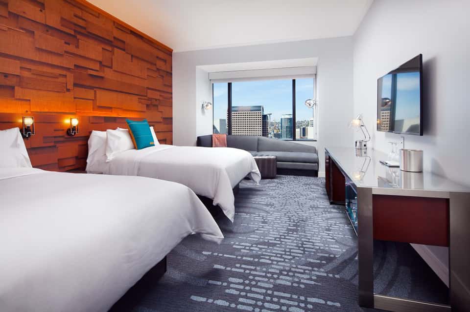 Modern hotel bedroom with two queen beds, wooden accent wall, city skyline views, and contemporary furnishings