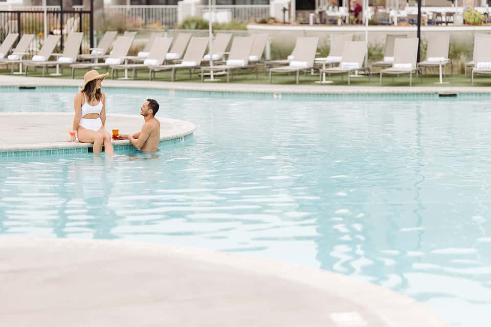 Couple relaxing at pool edge with lounge chairs and water view in background
