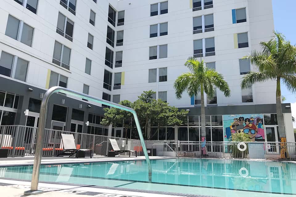 Modern white hotel building with turquoise pool, lounge chairs, and palm trees in courtyard