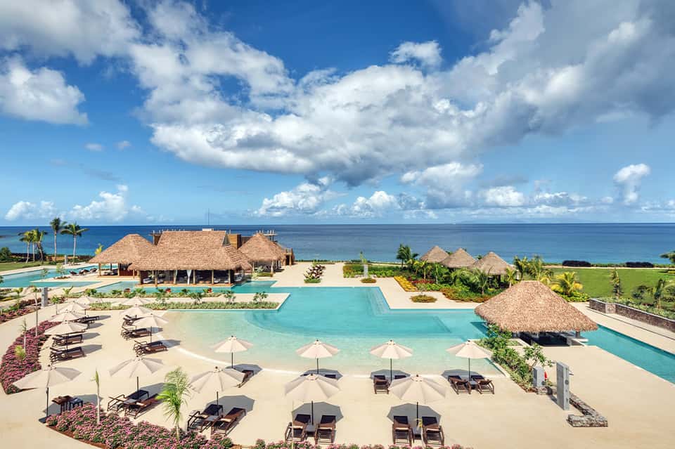 Luxury beachfront resort with turquoise pools, thatched-roof structures, white umbrellas, and ocean views