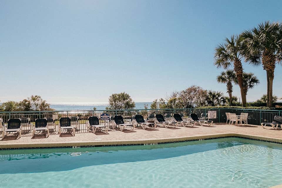 Resort pool deck with lounge chairs overlooking ocean and palm trees under clear blue sky