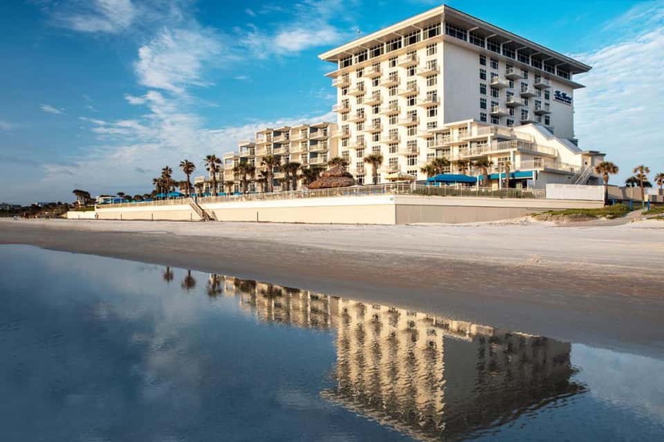 Beachfront resort hotel with multi-story white building and palm trees reflected in wet sand