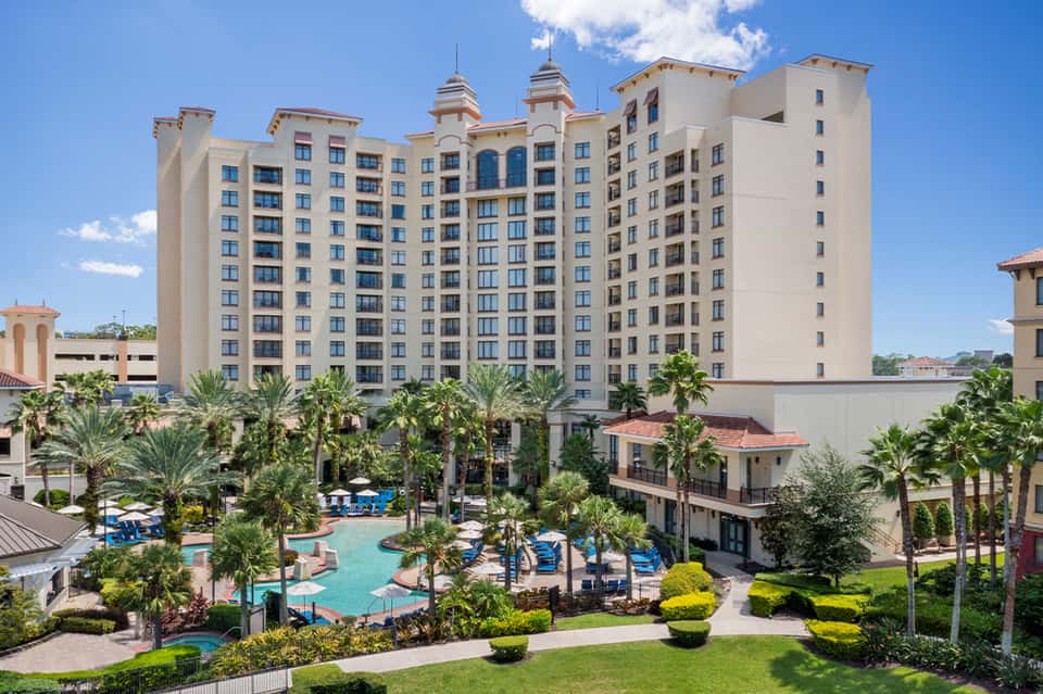 Large beige resort building with palm trees and curved swimming pool surrounded by landscaped grounds