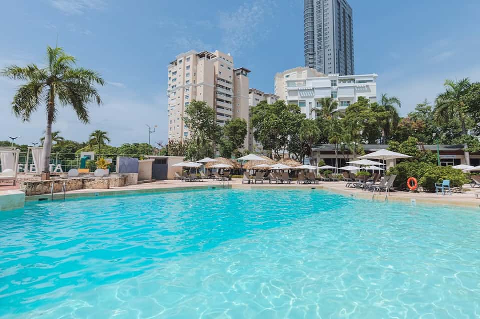 Turquoise lagoon-style pool with beach area, palm trees, and multi-story resort buildings under clear sky