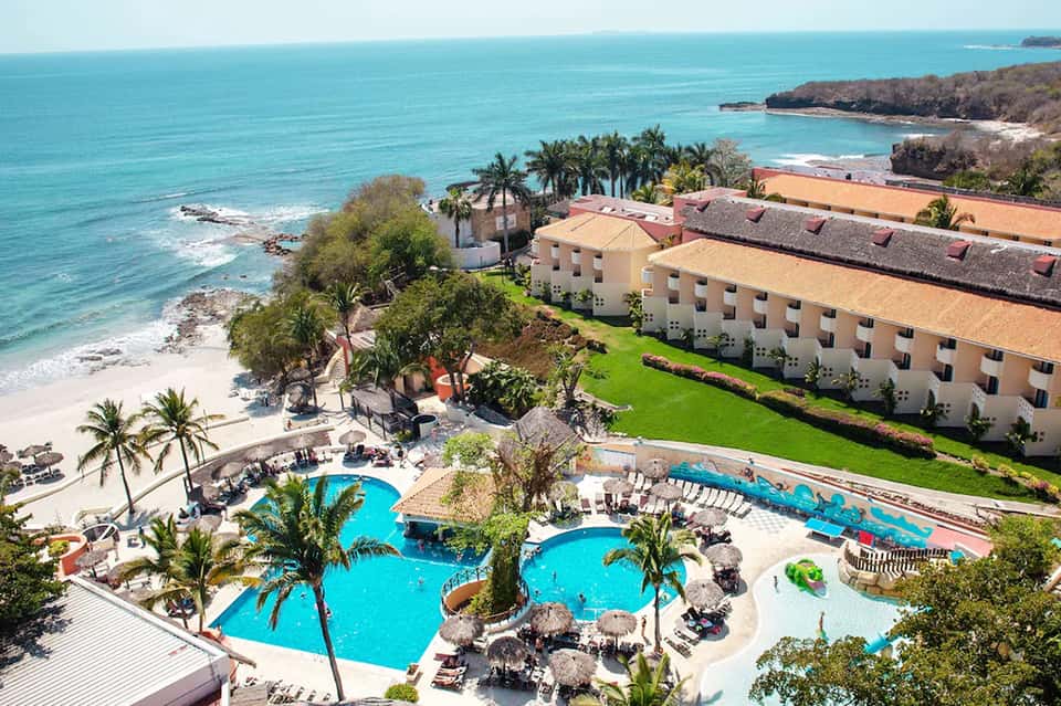 Beachfront resort with turquoise pools, palm trees, and tan buildings overlooking sandy beach and ocean