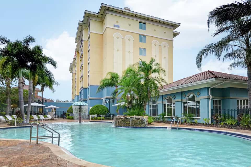 Yellow hotel building with pool, cabanas, and blue poolside structure surrounded by palm trees