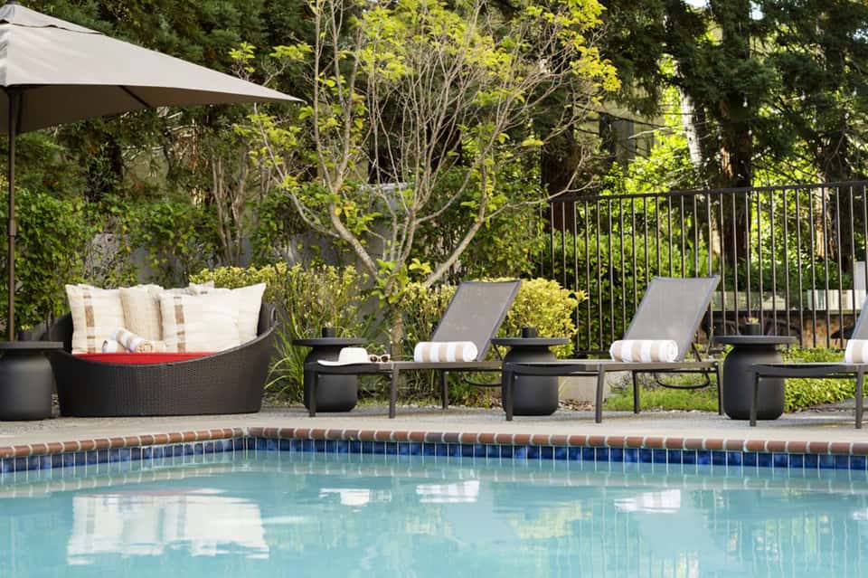Luxury poolside lounge area with black loungers, rattan seating, umbrella, and lush greenery surrounding a turquoise pool