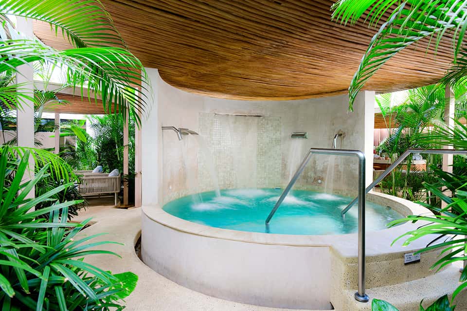 Indoor spa area with large circular hot tub surrounded by tropical plants and wooden ceiling