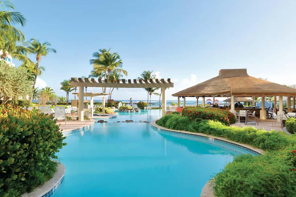 Tropical beachfront resort pool with turquoise water, tiki bar, palm trees, and ocean views