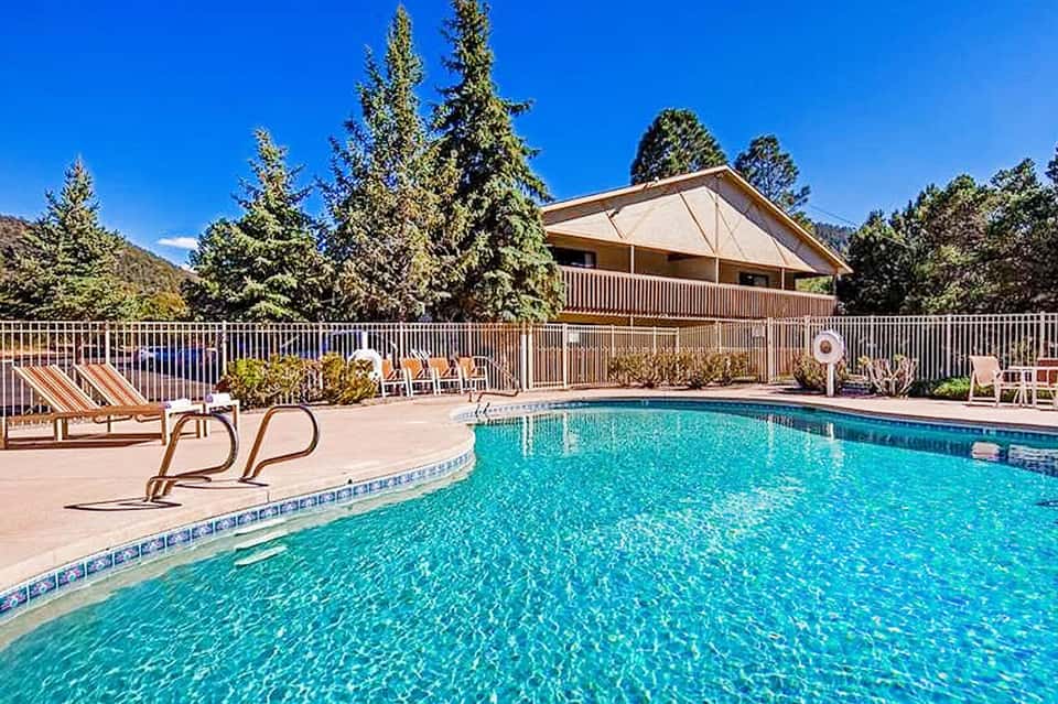Resort swimming pool with crystal-clear water, lounge chairs, and pine trees surrounding a tan building under blue sky