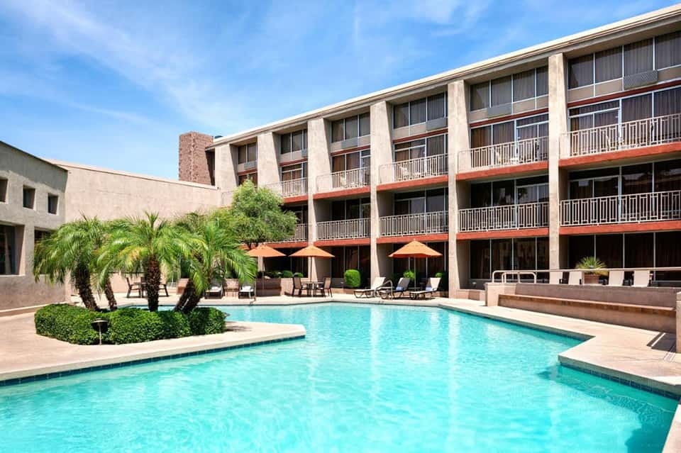 Resort courtyard with turquoise swimming pool, palm trees, orange umbrellas, and multi-story building with balconies