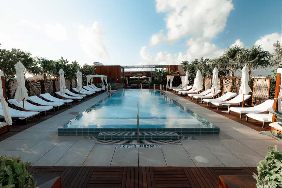 Resort pool deck with lap pool, white loungers, umbrellas, and palm trees under blue sky