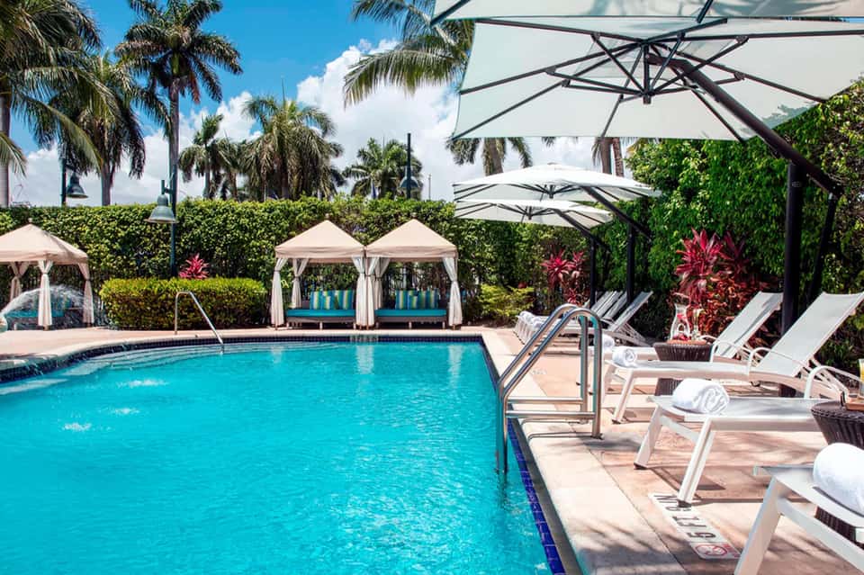 Resort pool with turquoise water, white cabanas, large umbrellas, and tropical palm landscaping