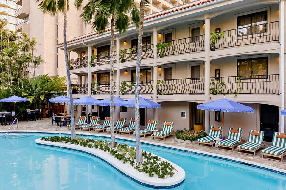 Multi-story hotel building with white columns, blue umbrellas, and striped lounge chairs by curved pool
