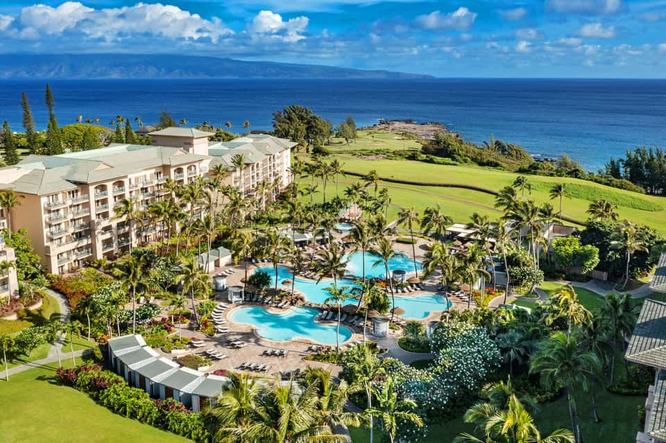 Aerial view of oceanfront resort with curved pools, palm trees, and manicured lawns overlooking blue ocean and distant island.