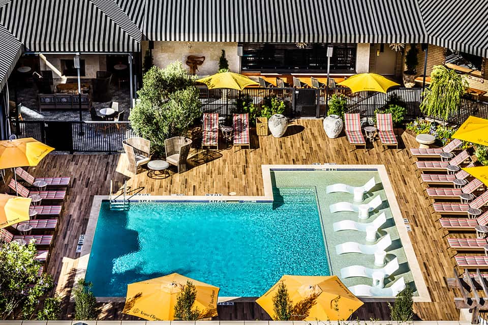 Aerial view of resort pool deck with yellow umbrellas, lounge chairs, and wooden decking surrounded by buildings