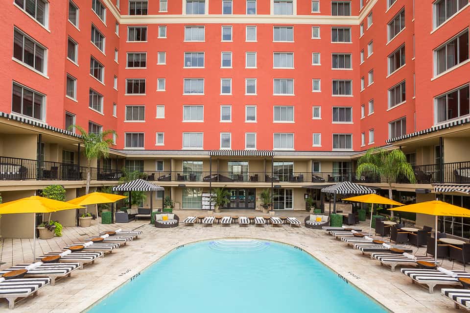 Coral-red courtyard building with striped lounge chairs, blue pool, yellow umbrellas, and black-and-white accent details