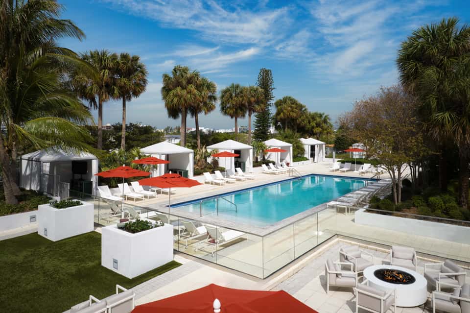 Resort pool with red umbrellas, white cabanas, lounge chairs, and manicured lawn with palm trees