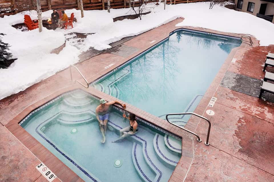 Guests enjoying hot tub in snow-covered resort courtyard with heated pool in background