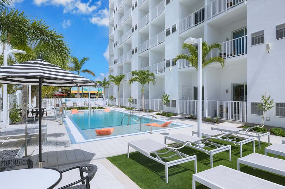 Modern white hotel building with blue pool, lounge chairs, and palm trees under clear sky
