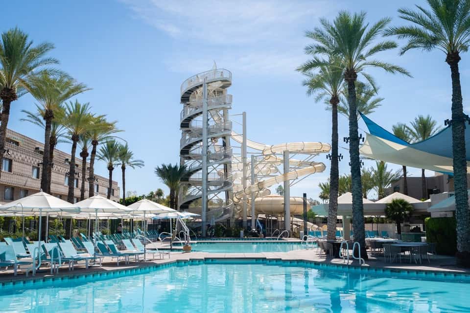 Resort pool with water slides, palm trees, lounge chairs, white umbrellas, and clear blue water under sunny skies