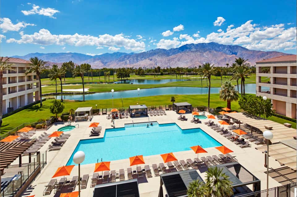 Luxury resort pool complex with mountain views, palm trees, and multiple lounging areas with orange umbrellas