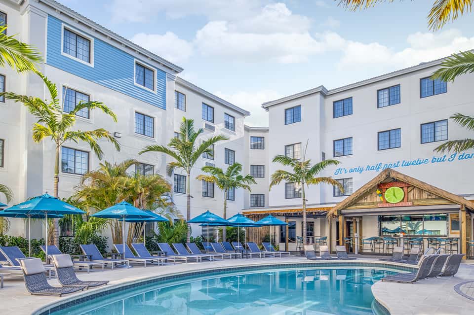 Resort pool with blue umbrellas and loungers, palm trees, hotel buildings with white walls