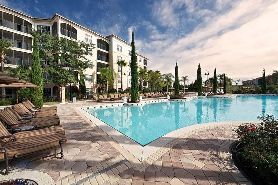 Resort pool with loungers, cypress trees, and multi-story hotel building under blue sky