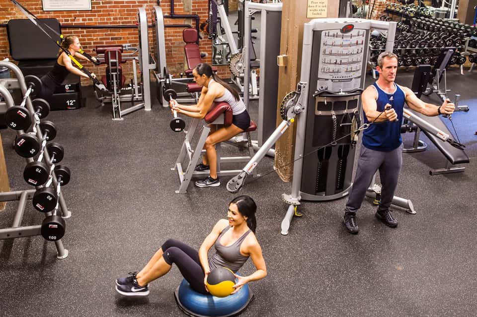 Indoor gym with fitness equipment, dumbbells, machines, and people exercising in brick-walled facility