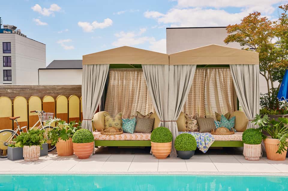 Rooftop poolside cabanas with beige canopies, white curtains, cushioned seating, potted plants, and city building views