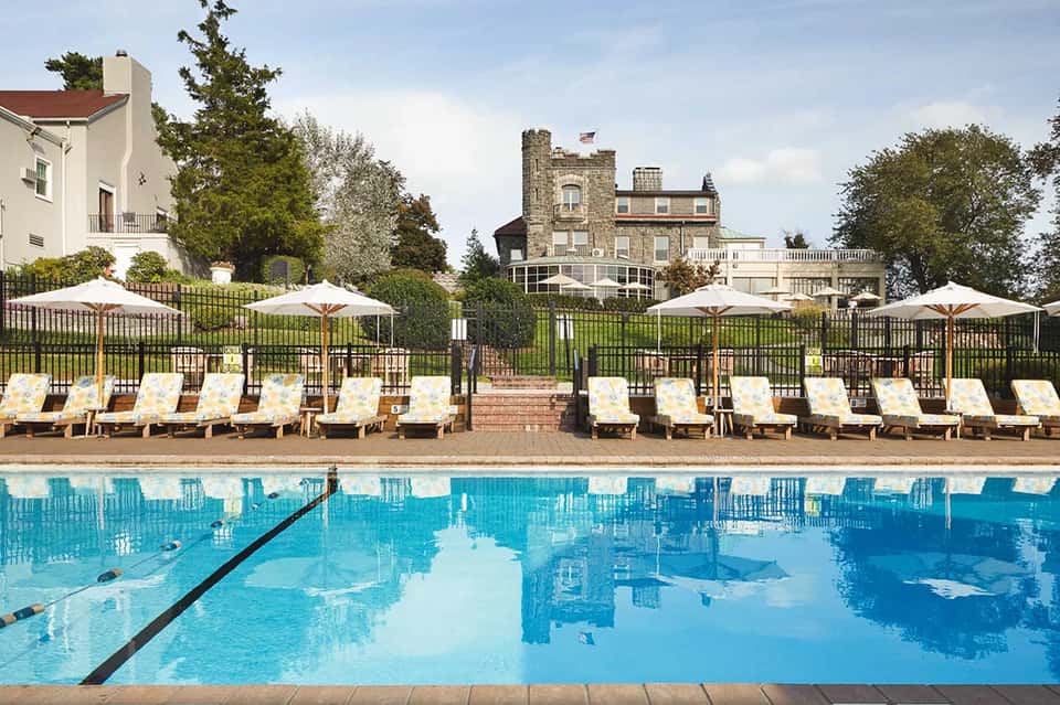 Outdoor swimming pool with lounge chairs and umbrellas, historic mansion visible beyond black fence