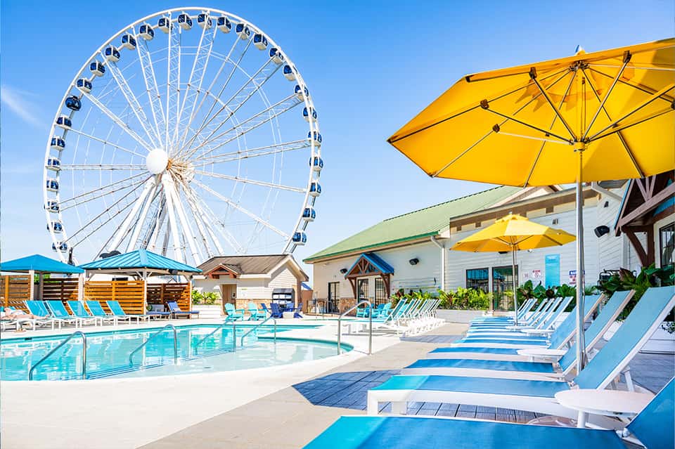 Resort pool area with Ferris wheel in background, yellow umbrellas, and blue lounge chairs