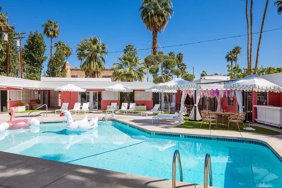 Retro-style resort pool with pink cabanas, white umbrellas, and palm trees under clear blue sky