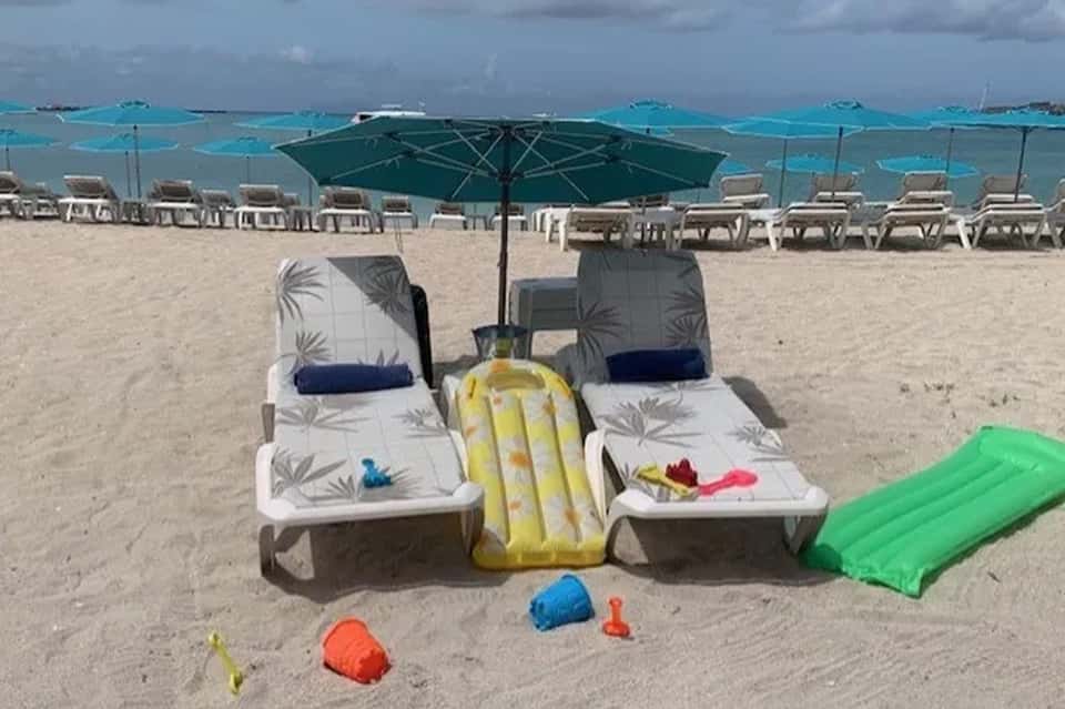 Beach setup with two lounge chairs under green umbrella, yellow float, and colorful beach toys on sand