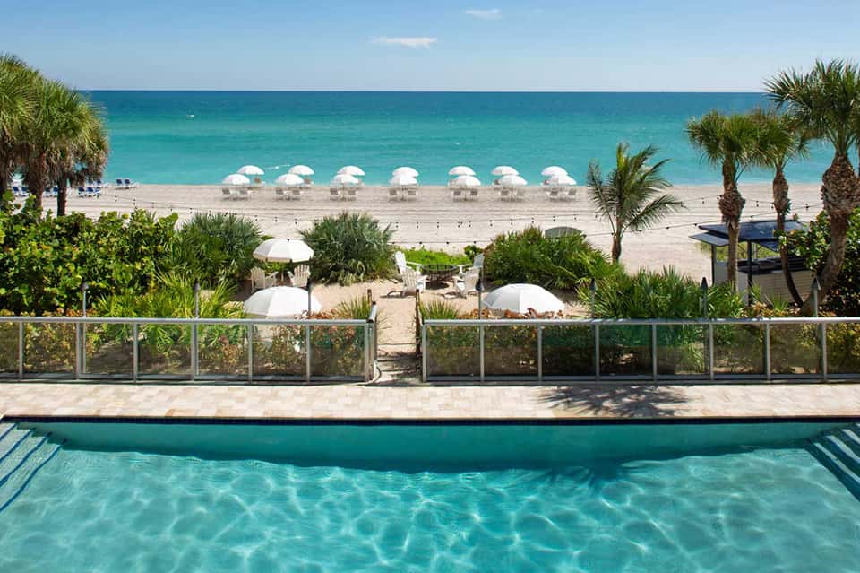 Resort pool with turquoise water overlooking pristine beach with white umbrellas and palm trees