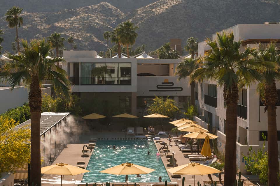 Resort courtyard with rectangular pool, yellow umbrellas, palm trees, and modern white architecture