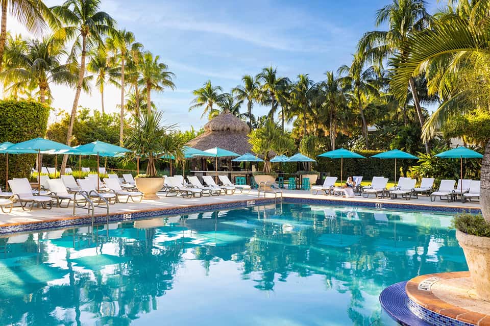 Resort pool surrounded by turquoise umbrellas, palm trees, and lounge chairs under blue sky