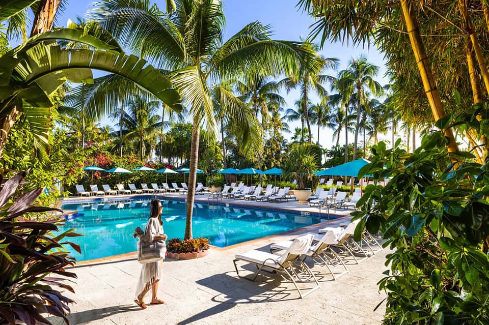 Tropical resort pool surrounded by palm trees, lounge chairs, and blue umbrellas under clear sky