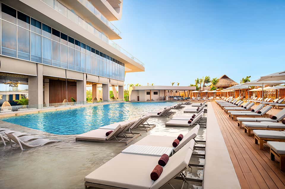 Resort pool deck with rows of cushioned lounge chairs, indoor swimming pool, and modern building architecture