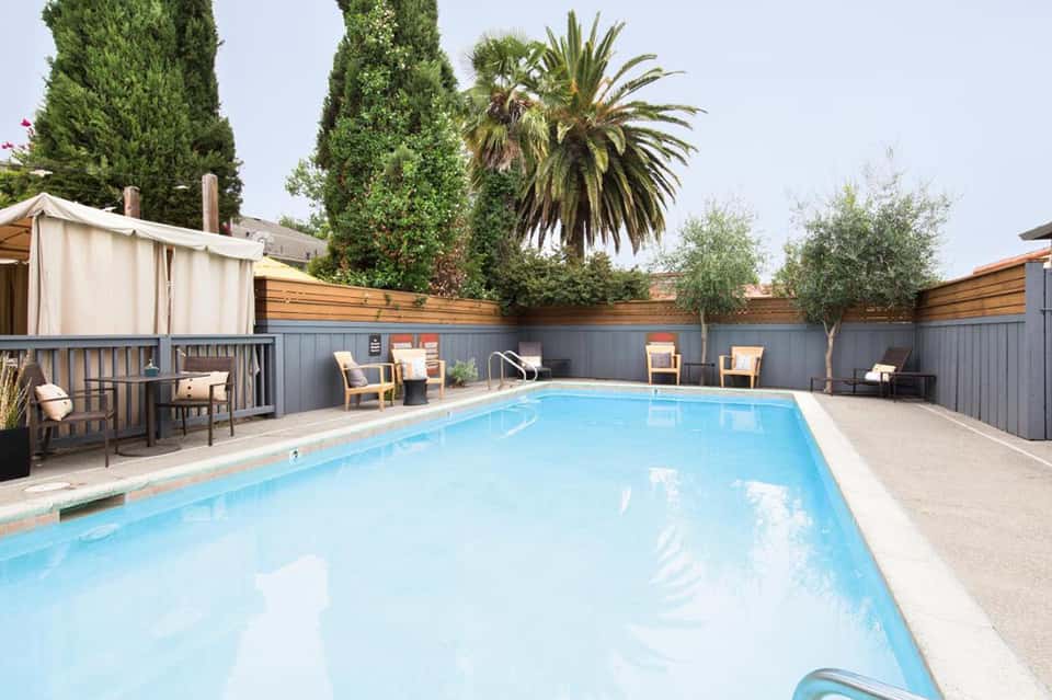 Resort swimming pool with wooden deck enclosed by gray fence, surrounded by lounge chairs and palm trees