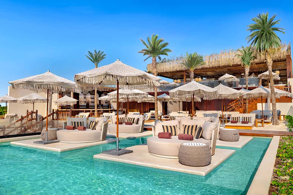 Luxurious beach club with fringe umbrellas, lounging pods, palm trees, and wooden structures on turquoise water