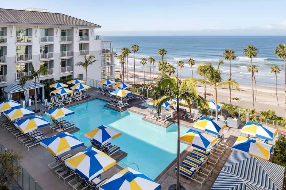 Beachfront resort pool with blue and yellow umbrellas, ocean view, and palm tree-lined shore