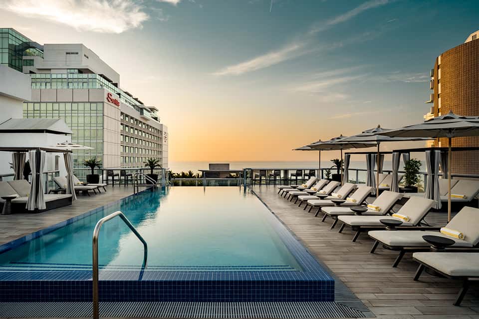 Rooftop infinity pool at sunset with loungers, umbrellas, and city skyline views