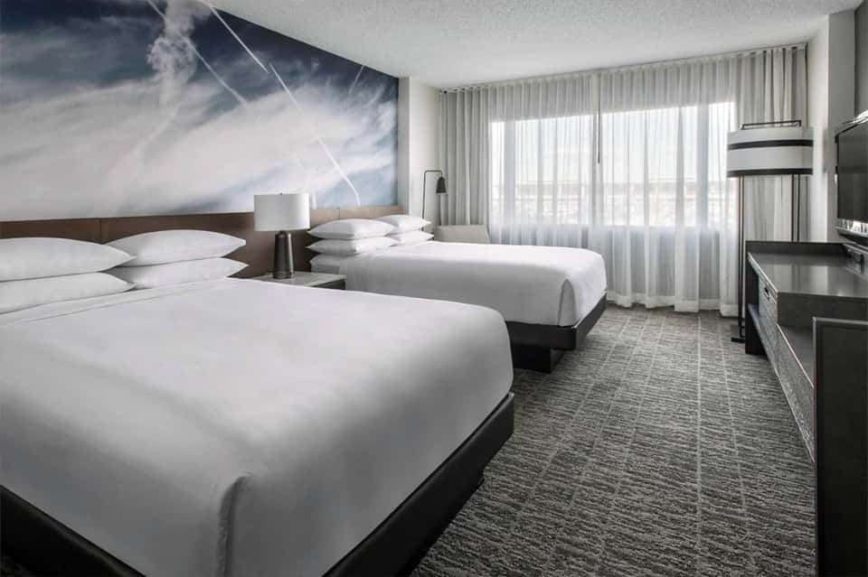 Modern hotel room with two queen beds, floor-to-ceiling windows, and cloud artwork on accent wall