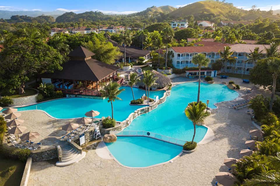 Resort pool complex with turquoise water, palm trees, tropical landscaping, and multi-story building