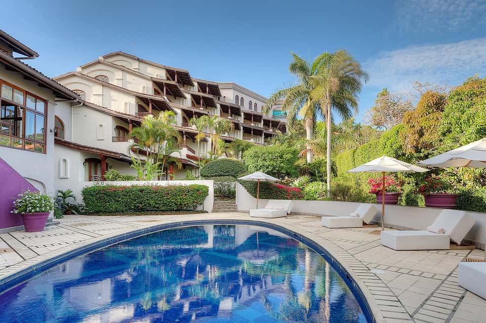 Luxury resort courtyard with pool, palm trees, white lounge chairs, umbrellas, and Spanish-style architecture