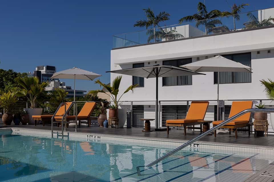 Resort poolside area with orange lounge chairs, gray umbrellas, palm trees, and modern white building