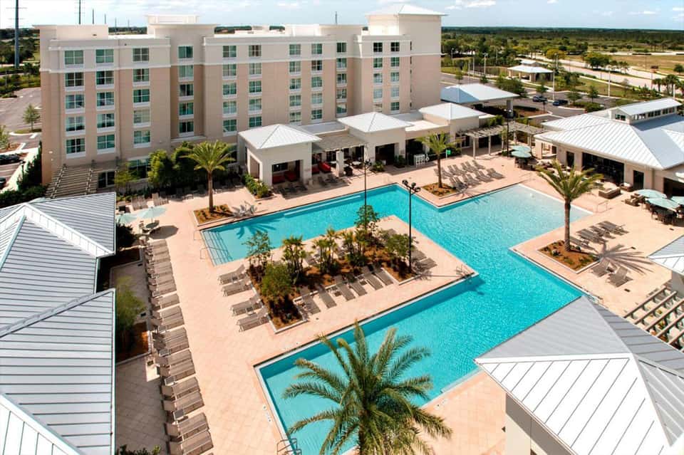 Aerial view of resort with multi-story hotel building, expansive pools, and surrounding palm trees