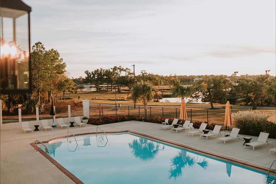 Lakeside pool at dusk with white lounge chairs, umbrellas, fence railing, and trees reflected in calm water