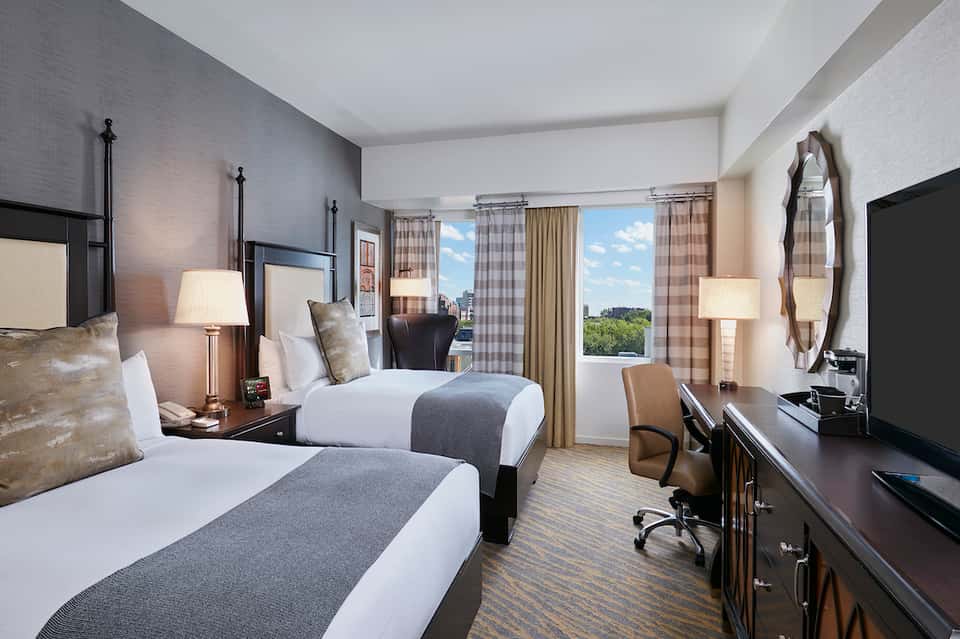 Modern hotel guest room with two queen beds, black metal frames, gray bedding, plaid curtains, and large windows overlooking grounds